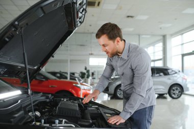 Looks under the hood of automobile. Young man in the car dealership