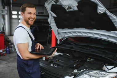 Otomobil servisi, tamirat, bakım konsepti. Tamirci arabayı kontrol ediyor, benzin istasyonunda dizüstü bilgisayarla tarama yapıyor. Endüstriyel motor tamiri hizmeti