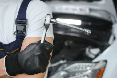 With tool in hand. Adult man in colored uniform works in the automobile salon.