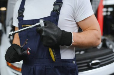With tool in hand. Adult man in colored uniform works in the automobile salon.