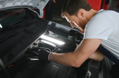A mechanic is doing car checkup under the hood with flashlight at mechanic's shop