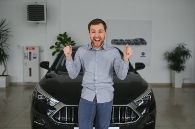 Emotional handsome guy raising hand up and smiling at camera. Rich man buying auto at luxury dealership salon.