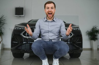 Emotional handsome guy raising hand up and smiling at camera. Rich man buying auto at luxury dealership salon.