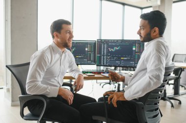 Two successful trader sitting in office and looking at screen, checking cryptocurrency information data on finance market graph, pointing on monitor.