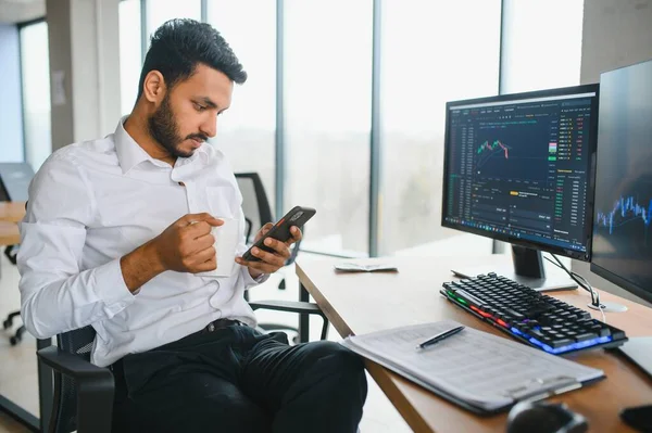 Side view of smart Indian crypto trader, investor, analyst broker, using laptop and smartphone analyzing digital cryptocurrency exchange, stock market charts, thinking of investing and funds risks.