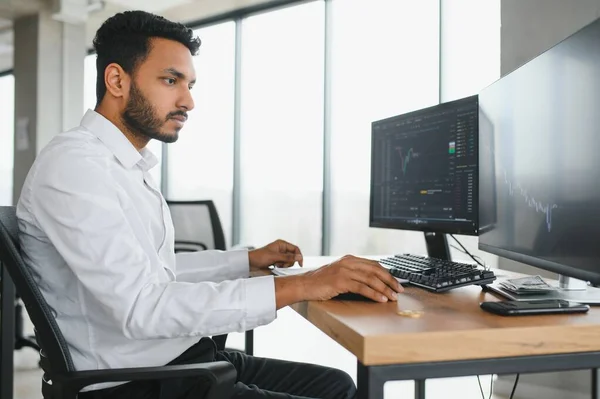 Young indian business man trader looking at computer screen with trading charts reflecting in eyeglasses watching stock trading market financial data growth concept, close up