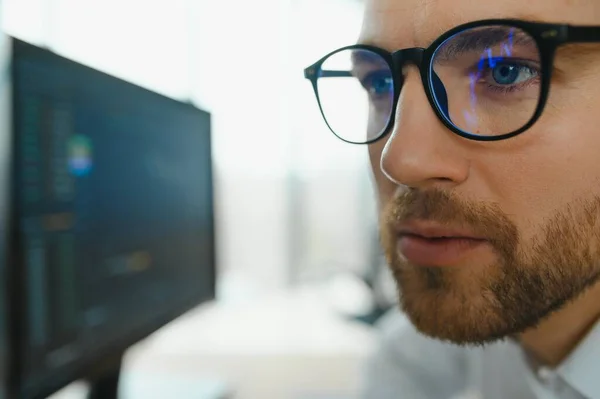 Young business man trader wearing glasses looking at computer screen with trading charts reflecting in eyeglasses watching stock trading market financial data growth concept, close up