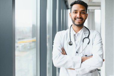 Modern özel klinikte kameraya bakarak duran, boynundaki steteskopla beyaz önlük giyen mutlu Hintli erkek doktor portresi. Tıbbi sağlık hizmeti kavramı.