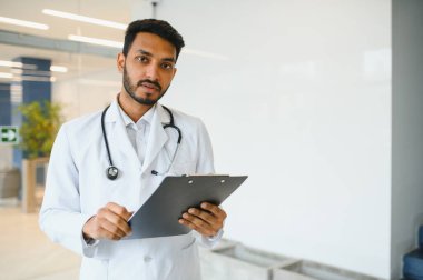 Modern özel klinikte kameraya bakarak duran, boynundaki steteskopla beyaz önlük giyen mutlu Hintli erkek doktor portresi. Tıbbi sağlık hizmeti kavramı.