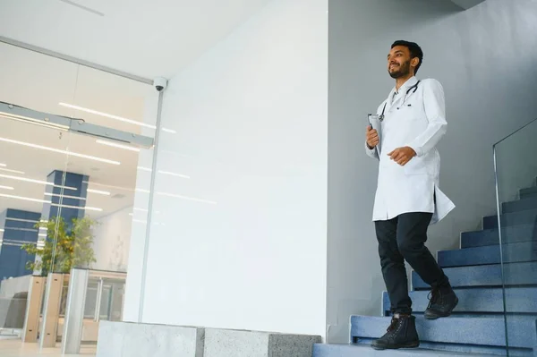 Portrait of male indian doctor with serious expression and crossed arms wearing white coat having open door on clinic corridor as background
