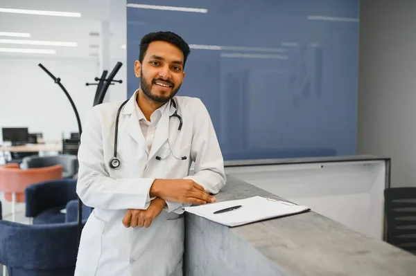 Portrait of male indian doctor with serious expression and crossed arms wearing white coat having open door on clinic corridor as background