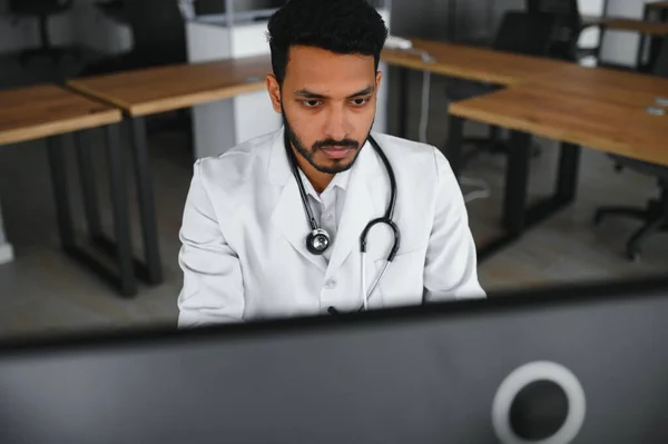 Indian male doctor physician wearing glasses, white medical gown and stethoscope sitting at the desk with the laptop in modern clinic and involved online video onference, consulting remotely