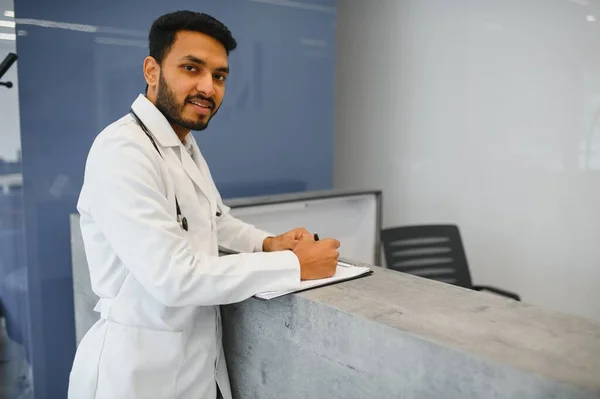 Portrait of happy friendly male Indian latin doctor medical worker wearing white coat with stethoscope around neck standing in modern private clinic looking at camera. Medical healthcare concept.