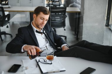 Fatigue young businessman sitting at workplace and holding whiskey bottle, drinking alcohol, bankruptcy concept
