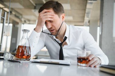 Fatigue young businessman sitting at workplace and holding whiskey bottle, drinking alcohol, bankruptcy concept