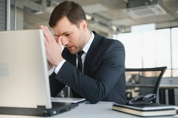Young stressed handsome businessman working at desk in modern office shouting at laptop screen and being angry about financial situation, jealous of rival capabilities, unable to meet client needs.