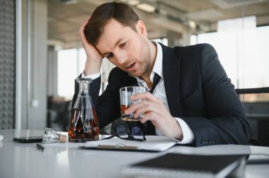 Fatigue young businessman sitting at workplace and holding whiskey bottle, drinking alcohol, bankruptcy concept