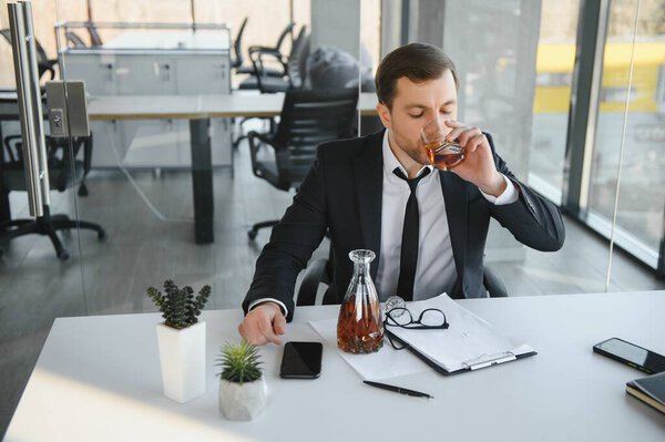Fatigue young businessman sitting at workplace and holding whiskey bottle, drinking alcohol, bankruptcy concept