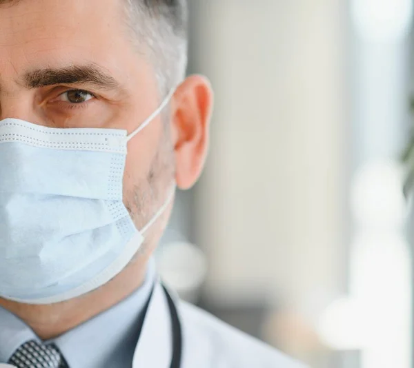 Mature old medical healthcare professional doctor wearing white coat, stethoscope, glasses and face mask standing in hospital. Medical staff health care protection concept. Portrait.