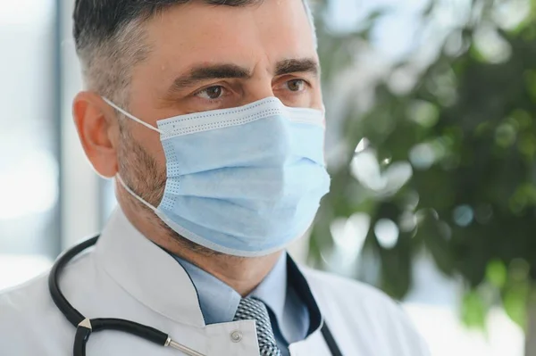 Mature old medical healthcare professional doctor wearing white coat, stethoscope, glasses and face mask standing in hospital. Medical staff health care protection concept. Portrait.