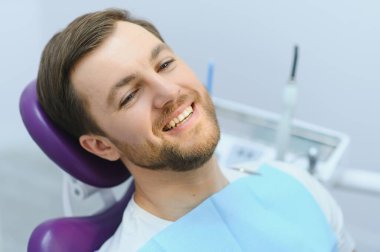 Diş muayenesi sırasında gülümseyen yakışıklı bir adam. Mutlu erkek hasta dişçi koltuğunda oturup dişlerini kontrol ediyor.