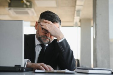 Koyu renk takım elbiseli endişeli işadamı, kitap ve evraklarla dolu ofis masasında oturarak iş lerle aşırı yükleniyor