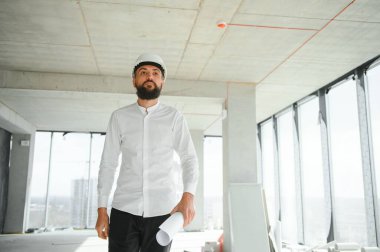 Serious busy young attractive arabic industrial engineer in hard hat with project drawings in factory interior. Professional and industry, builder architect job, worker checking work at plant indoor.