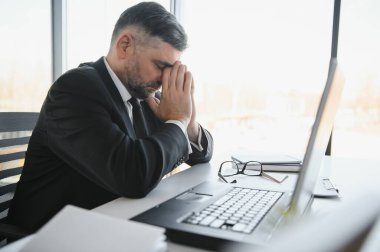 Koyu renk takım elbiseli endişeli işadamı, kitap ve evraklarla dolu ofis masasında oturarak iş lerle aşırı yükleniyor