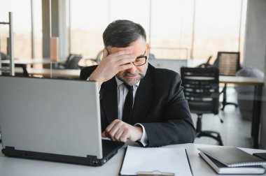 Koyu renk takım elbiseli endişeli işadamı, kitap ve evraklarla dolu ofis masasında oturarak iş lerle aşırı yükleniyor