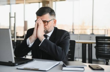 Koyu renk takım elbiseli endişeli işadamı, kitap ve evraklarla dolu ofis masasında oturarak iş lerle aşırı yükleniyor