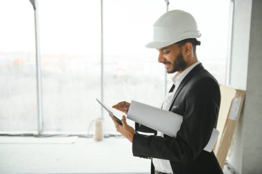 Kendine güvenen Asyalı mühendis modern bina inşaatında kontrol ve onarım için tablet kullanıyor. İnşaat alanında beyaz kaskla çalışan mimar