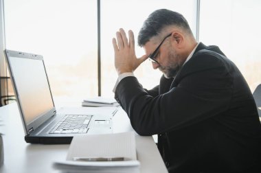 Koyu renk takım elbiseli endişeli işadamı, kitap ve evraklarla dolu ofis masasında oturarak iş lerle aşırı yükleniyor