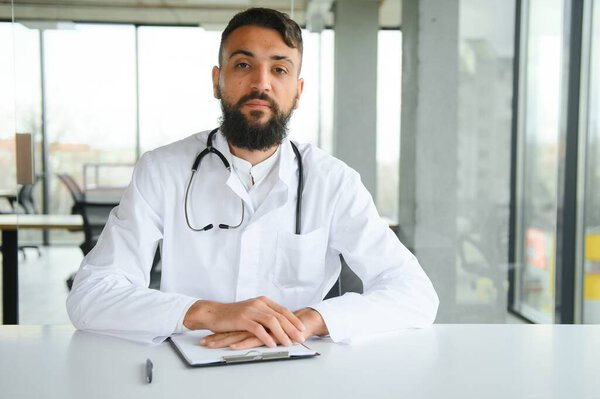 Young 30-aged handsome pleasant Arabic male doctor in white coat, posing at camera indoors