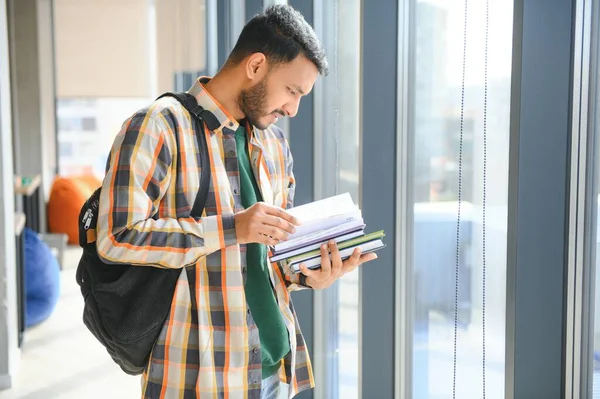 Young indian student boy reading book studying in college library with bookshelf behind. working on assignment or project.