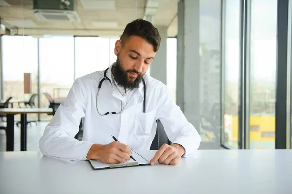 Portrait of arabic doctor handsome young man in workwear posing at modern clinic, sitting at workdesk.
