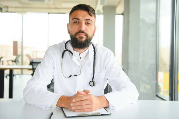 Young 30-aged handsome pleasant Arabic male doctor in white coat, posing at camera indoors