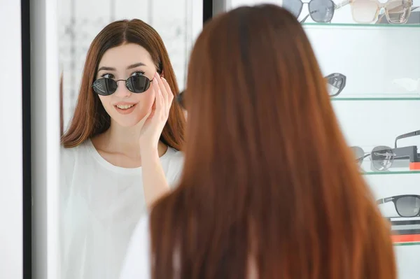 鏡の近くの店に立ってサングラスをしようとしている若い女性のショッピングの肖像画 — ストック写真