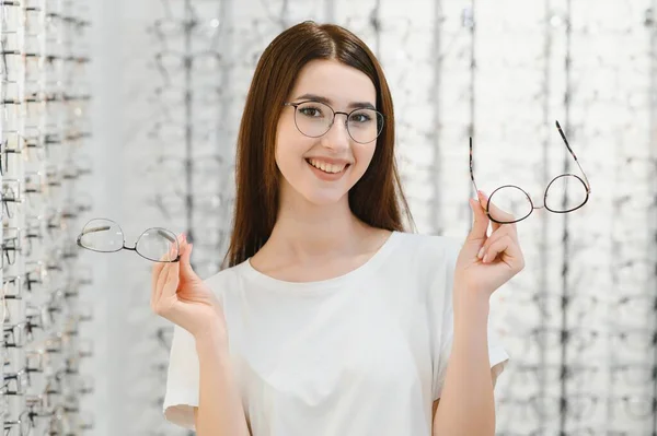 Health care, eyesight and vision concept - happy woman choosing glasses at optics store. Young beautiful girl ar optic store trying on new glasses.