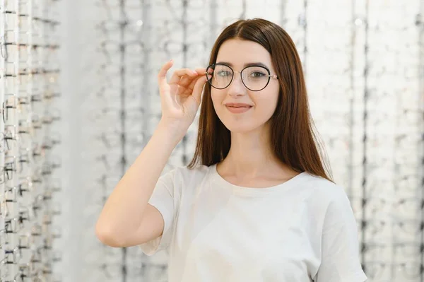 Health care, eyesight and vision concept - happy woman choosing glasses at optics store. Young beautiful girl ar optic store trying on new glasses.