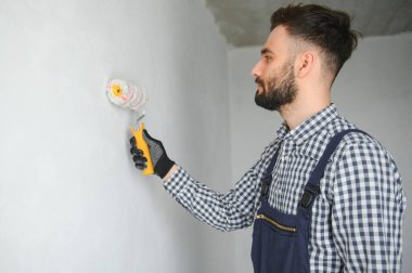 Mavi üniformalı genç, gülümseyen profesyonel işçi gri duvarları onarmak için yeni apartman dairesinde boya silindiriyle duruyor.