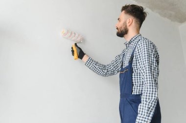 Mavi üniformalı genç, gülümseyen profesyonel işçi gri duvarları onarmak için yeni apartman dairesinde boya silindiriyle duruyor.