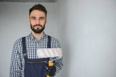 Mavi üniformalı genç, gülümseyen profesyonel işçi gri duvarları onarmak için yeni apartman dairesinde boya silindiriyle duruyor.