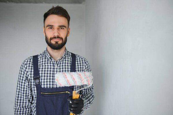 Young smiling professional worker in blue uniform standing with paint roller in new apartment for repairing over grey walls background, copy space