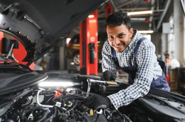 Hintli araba tamircisi benzin istasyonunda duruyor ve çalışıyor. Araba uzmanları kaldırılan arabayı inceliyor. Profesyonel tamirciler mavi üniforma giyiyor.