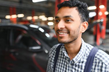 Hintli araba tamircisi benzin istasyonunda duruyor ve çalışıyor. Araba uzmanları kaldırılan arabayı inceliyor. Profesyonel tamirciler mavi üniforma giyiyor.