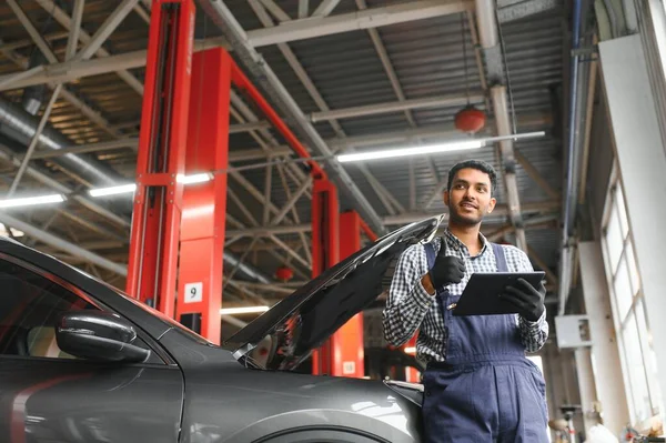 Indian car mechanic standing and working in service station. Car specialists examining the lifted car. Professional repairmen wearing mechanic uniform in blue color