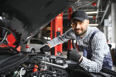 Profesyonel tamirci araba servisinde bir araba üzerinde çalışıyor. Güvenlik Gözlüklü Tamirci bir aracın motorunu tamir ediyor. Uzman bir Ratchet ile vidaları söküyor. Modern Temiz Atölye.