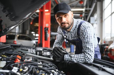 Profesyonel tamirci araba servisinde bir araba üzerinde çalışıyor. Güvenlik Gözlüklü Tamirci bir aracın motorunu tamir ediyor. Uzman bir Ratchet ile vidaları söküyor. Modern Temiz Atölye.