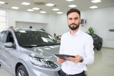 Otomobil galerisindeki resmi kıyafetler içinde elinde pano tutan, kendine güveni tam beyaz bir erkek dükkan asistanının yakın çekimi..
