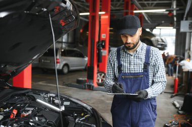 Araç motorunun yanında duran ve kaputu açık arızayı tespit eden profesyonel beyaz araba tamircisi..